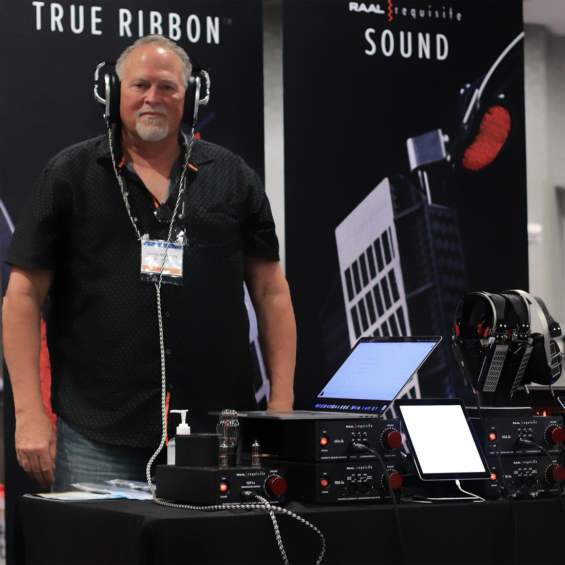 Danny of RAAL-requisite behind a trade show table featuring RAAL amplifiers and headphones