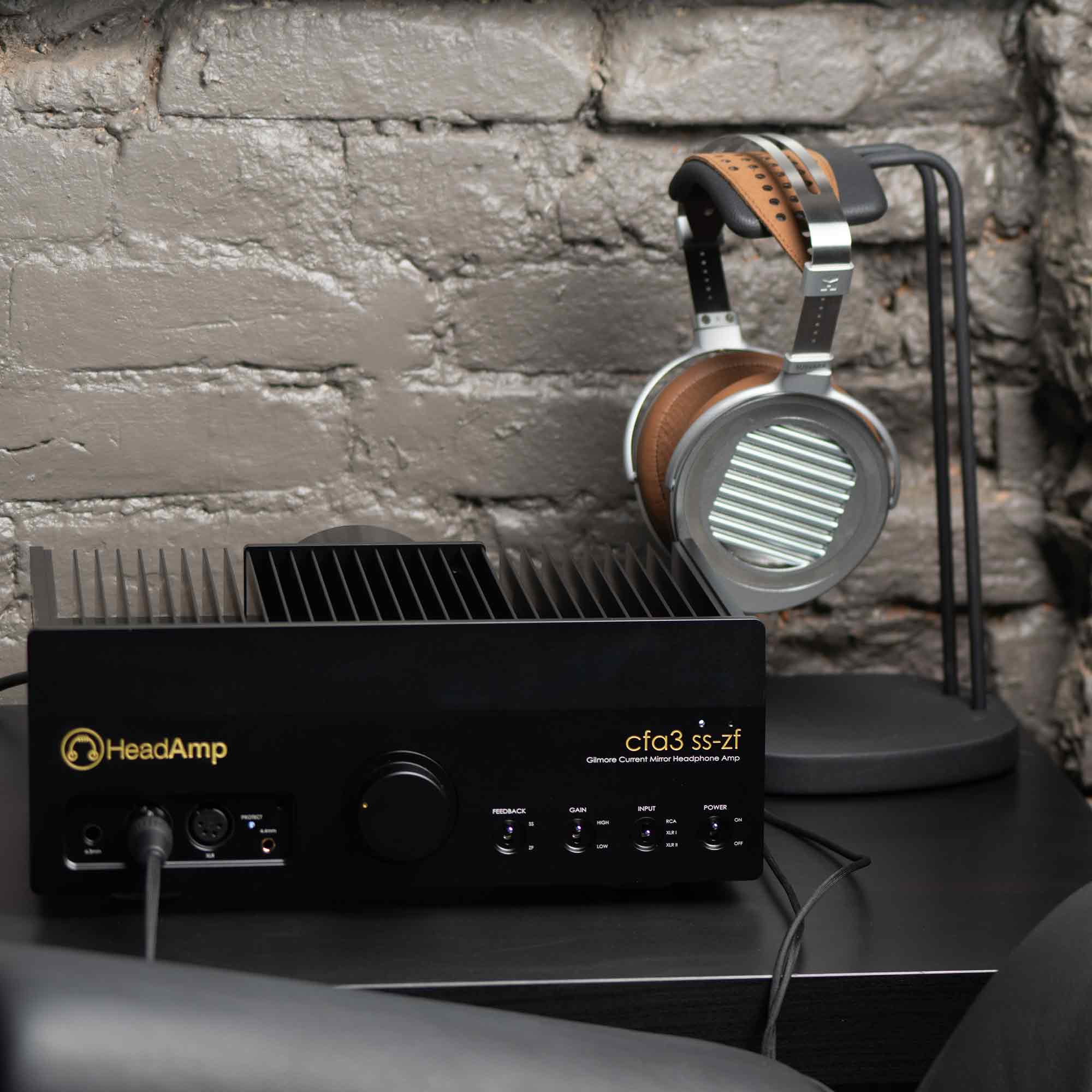 HeadAmp CFA3 reference headphone amp on black side table with HIFIMAN Susvara Unveiled headphones hanging on stand in front of grey brick wall