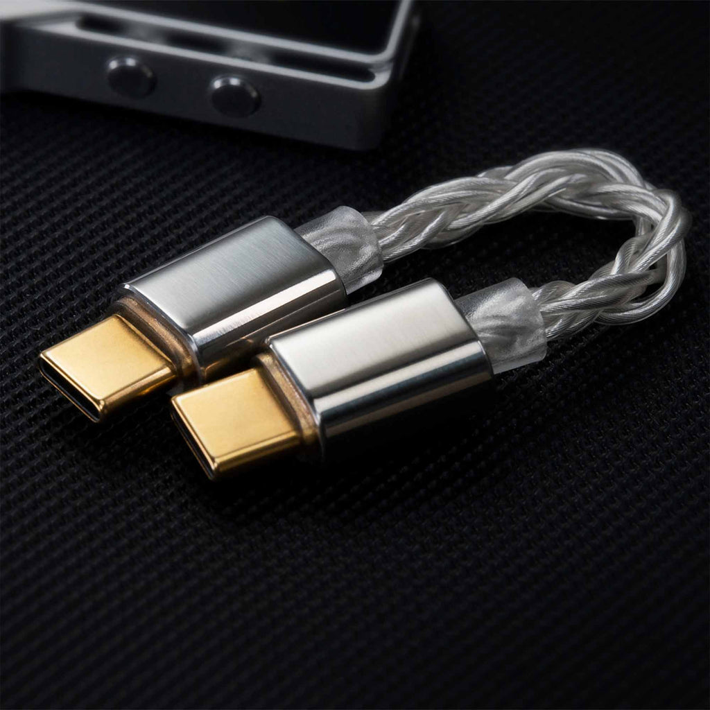 Two iBasso CB18 USB-C to USB-C OTG cables with metallic, gold-plated connectors and braided silver design lie side by side on a black textured surface, with part of an electronic device visible in the background.