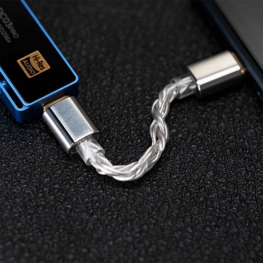 Close-up of the iBasso CB18 USB-C to USB-C OTG Cable, showing its braided design and metal connectors connected between a blue Hi-Res Audio device and a black electronic device on a textured black surface.