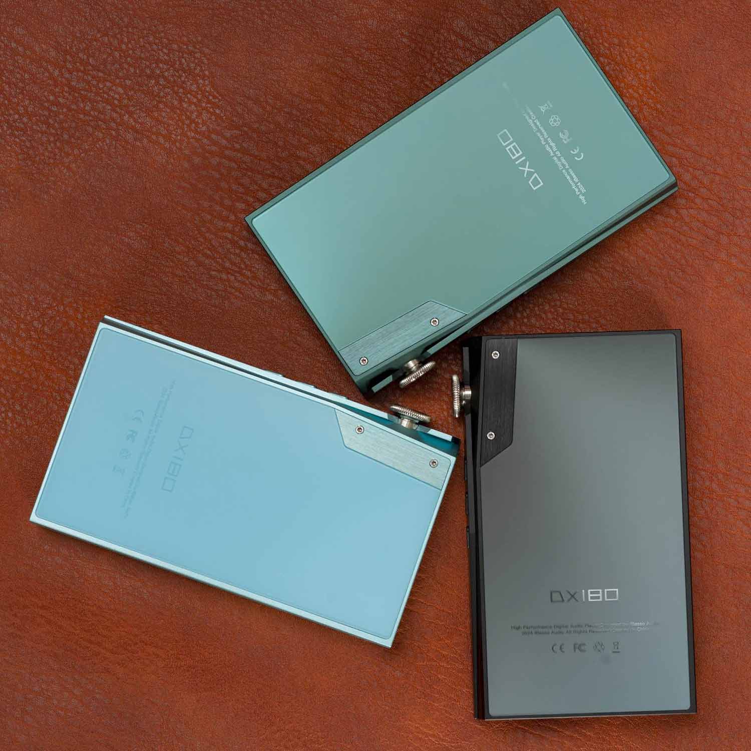 Three iBasso DX180 Digital Audio Players in black, green, and blue colors placed on brown leather surface