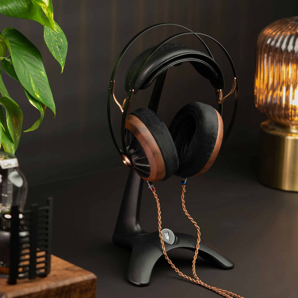 Meze Manta Headphone Stand holding wooden-cup headphones with braided cable on a dark desk near a lamp and plant