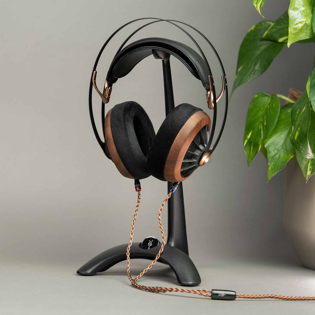 Meze Manta Headphone Stand holding wooden and black headphones with braided cable on gray surface near green plant