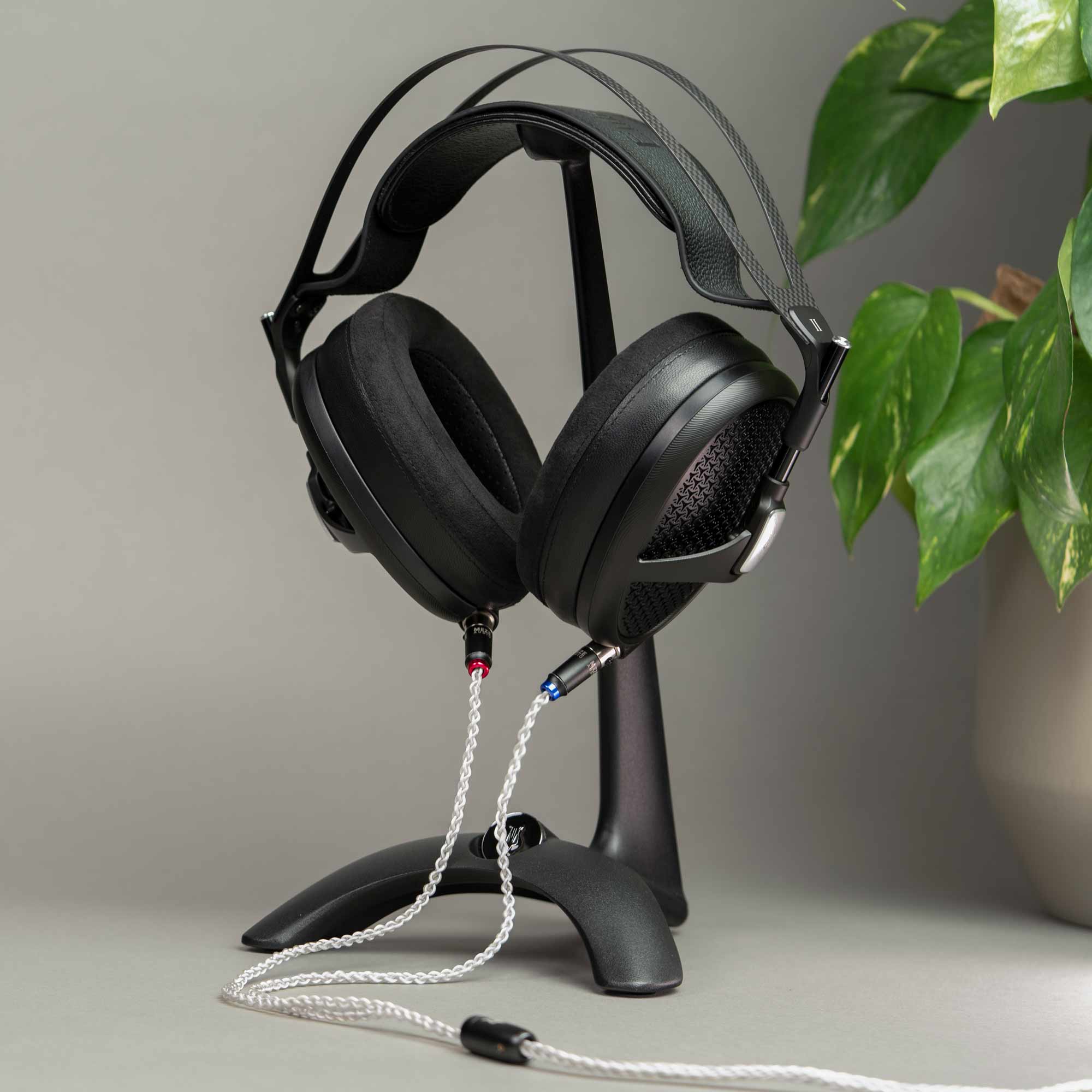 Meze Manta Headphone Stand holding black headphones with detachable braided cables on a gray surface near green plant