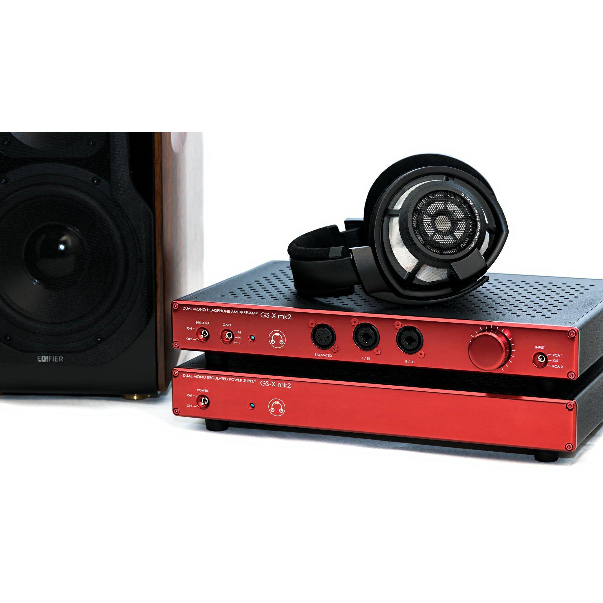 HeadAmp GS-X mk2 Balanced Headphone Amplifier / Pre-Amplifier in red with headphones placed on top and speaker in background