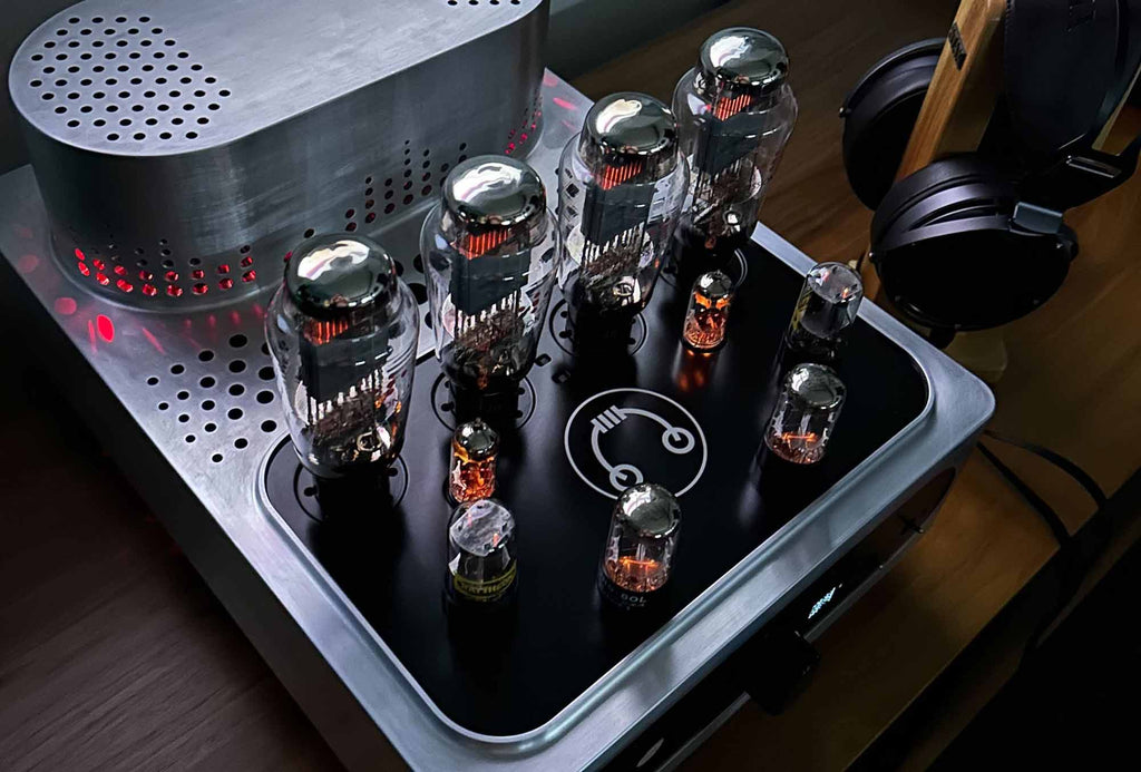 Close-up of HeadAmp Grand Cayman DHT Electrostatic Amp pre-order deposit showing glowing vacuum tubes and metal chassis on wooden surface
