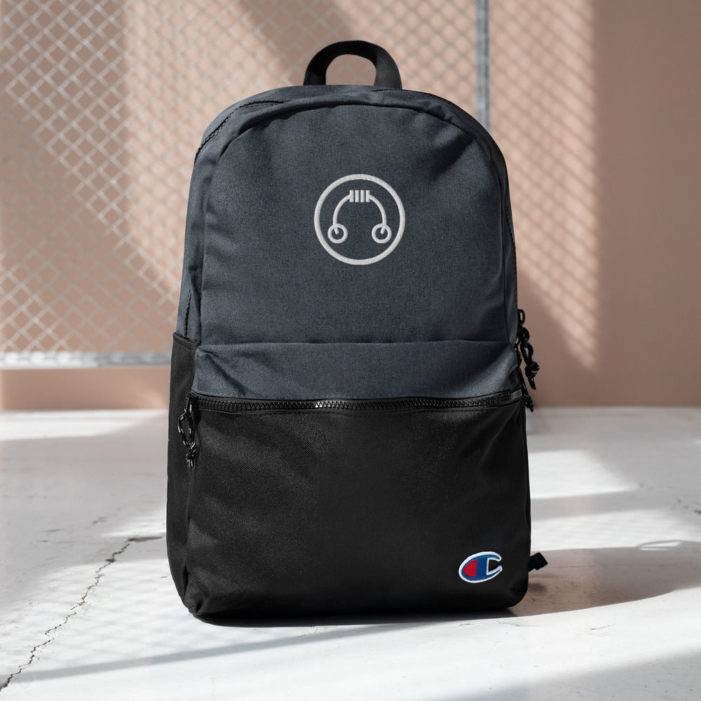 HeadAmp Embroidered Champion Backpack in black with front zipper pocket and embroidered logo on a lit concrete floor